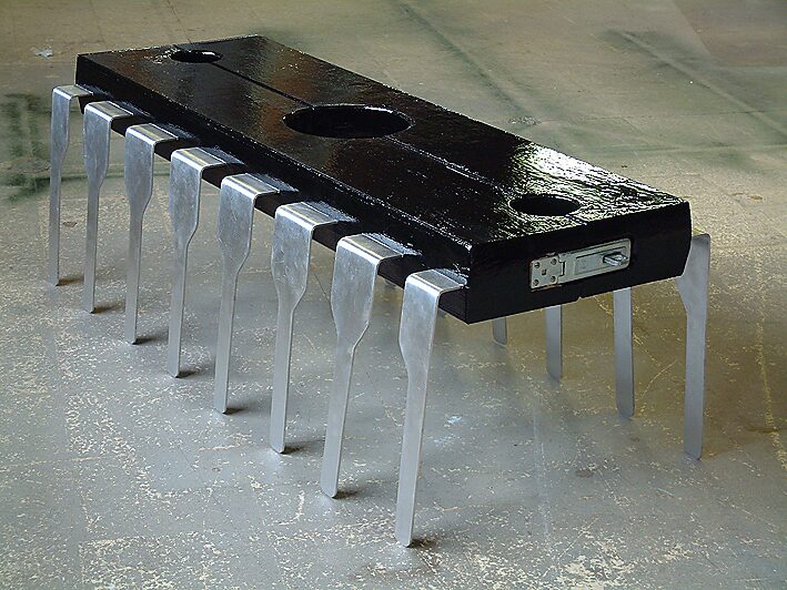 Digital Pillory — sculpture combining an enlarged integrated circuit (IC chip) with the form of a historical pillory clamp; a black wooden element with openings rests on repetitive aluminium legs referring to electronic contact pins.