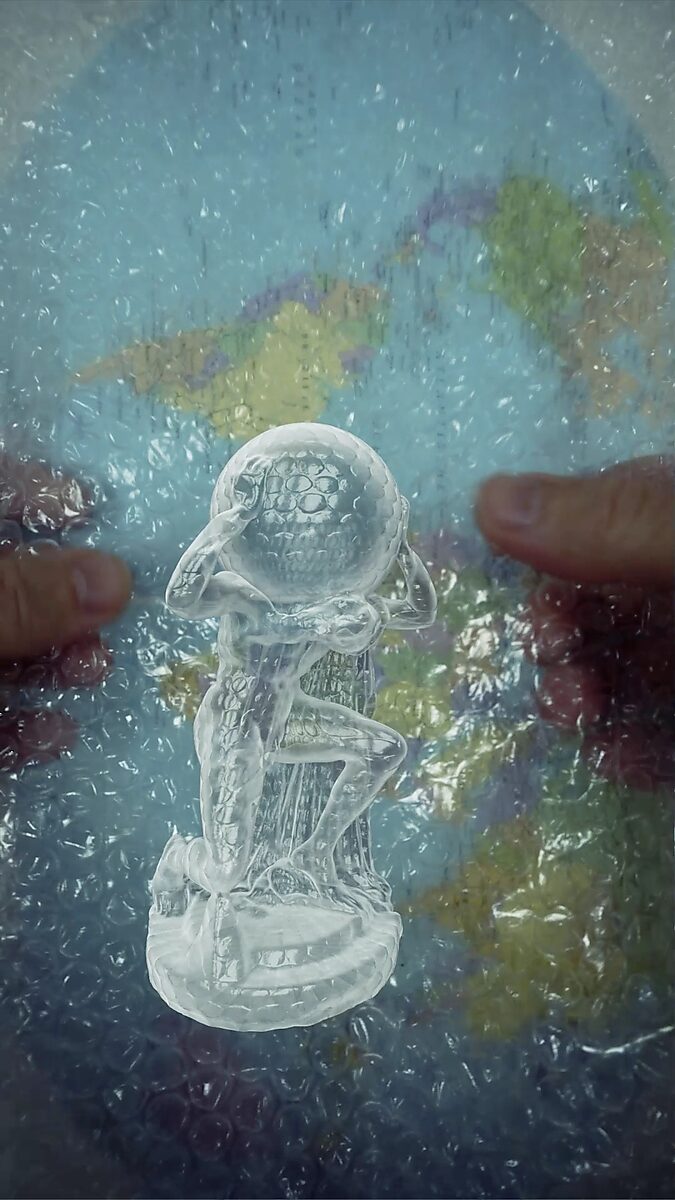 Virtual Transformation — Atlas in Bubble Wrap: video still from a 3D animation; a transparent Atlas-like figure holds a spherical form above as hands stretch bubble wrap over a world map in the background.