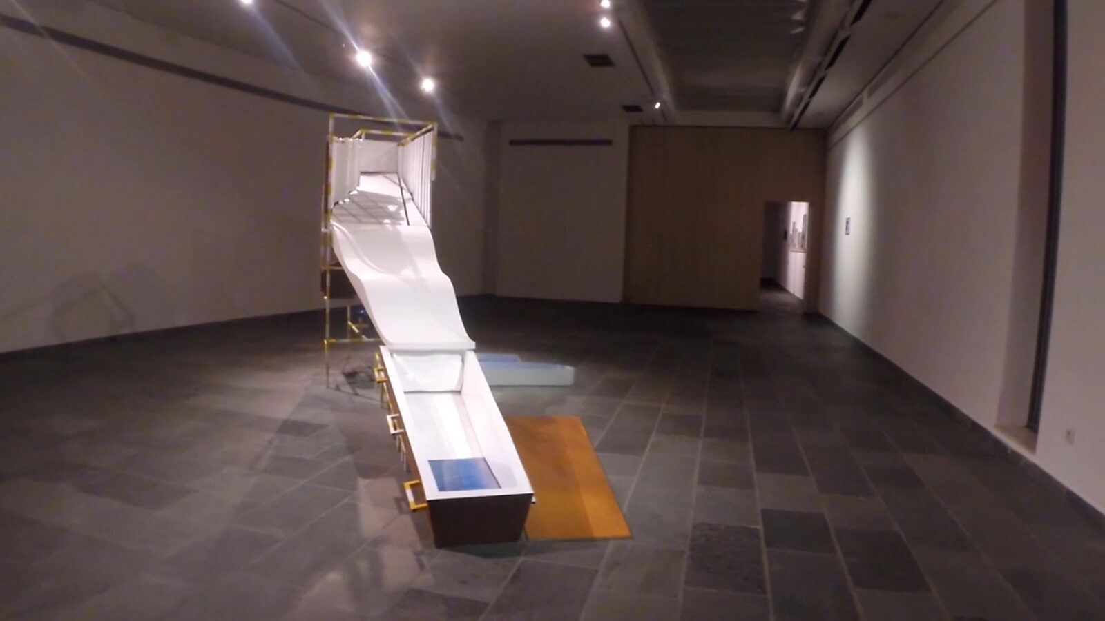 Constructie met blauw — videostill waarin het witte golvende polyester element overgaat in een neerwaartse schuifbeweging, waarbij de lineaire route zich versnelt richting het einde.