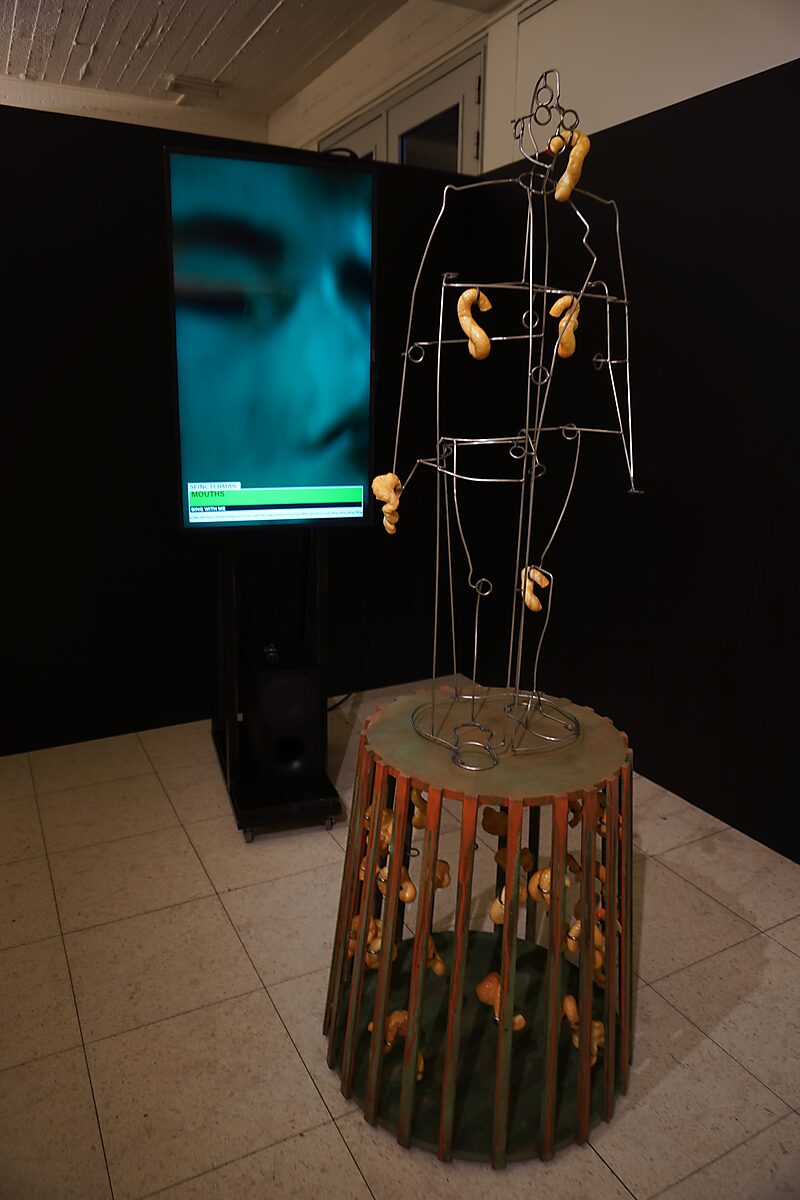 'Sfincterman for President’: Tentoonstellingszicht met draadfiguursculptuur met gele vormen op een cilindervormige sokkel; naast een verticaal videoscherm met close-up van een gezicht, Dommelhof, Pelt, 2024.
