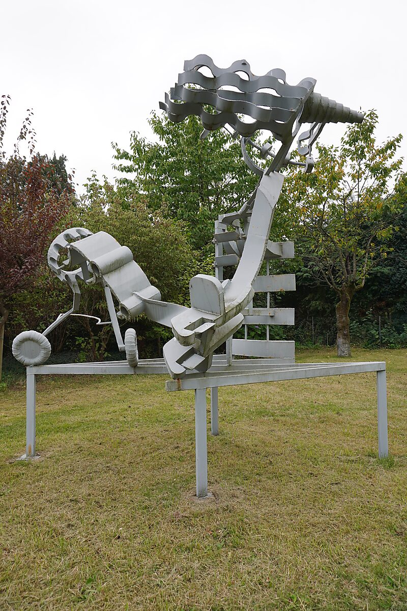 Abstractie van een vliegtuig — groot lichtgrijs sculptuur met gebogen en gelaagde metalen vormen die doen denken aan vleugels, turbines en cockpitstructuren, opgesteld op een verhoogd metalen frame in de tuin van de kunstenaar.
