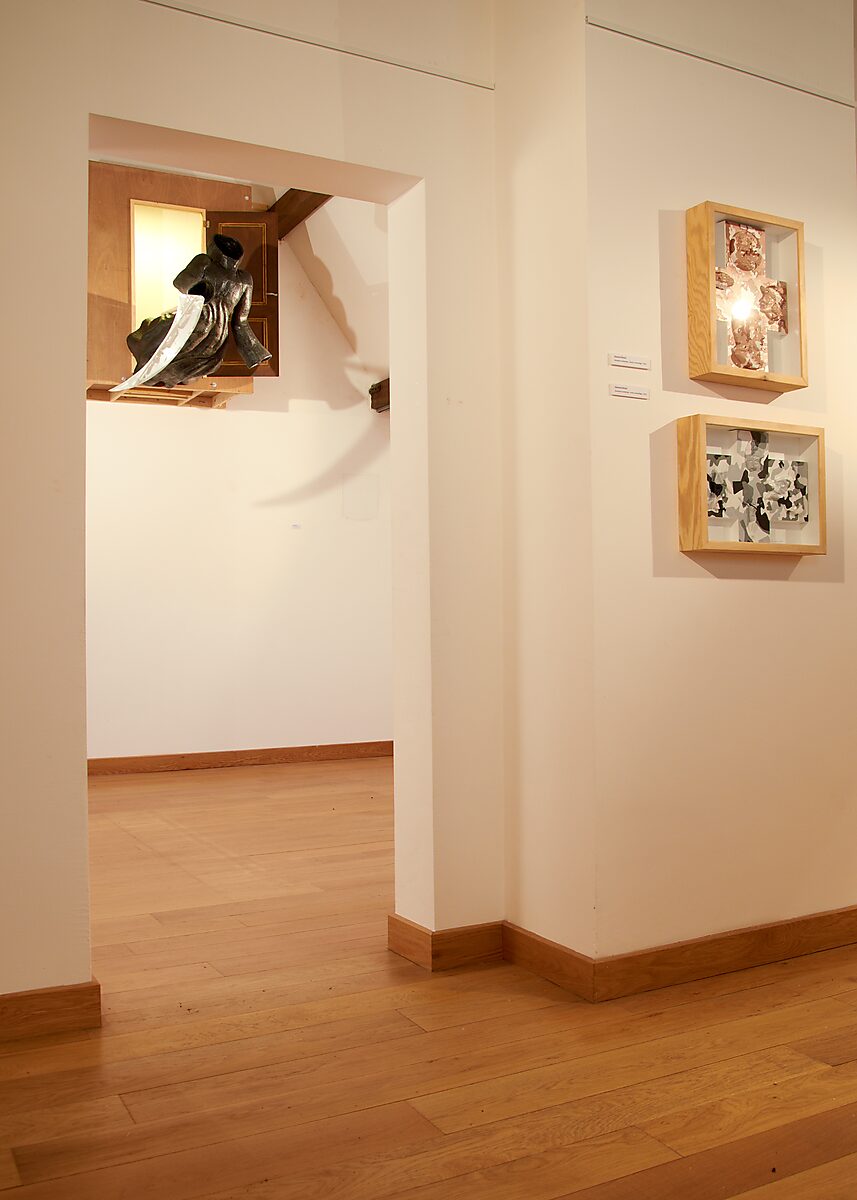 Duo expo met Raymond Minnen: Doorkijk naar wand met twee ingelijste reliëfwerken; in nis een sculptuur van een zwarte jasvorm die uit een open houten kast steekt en een lange steel met blad draagt, CC De Kimpel, Alden Biesen,2011