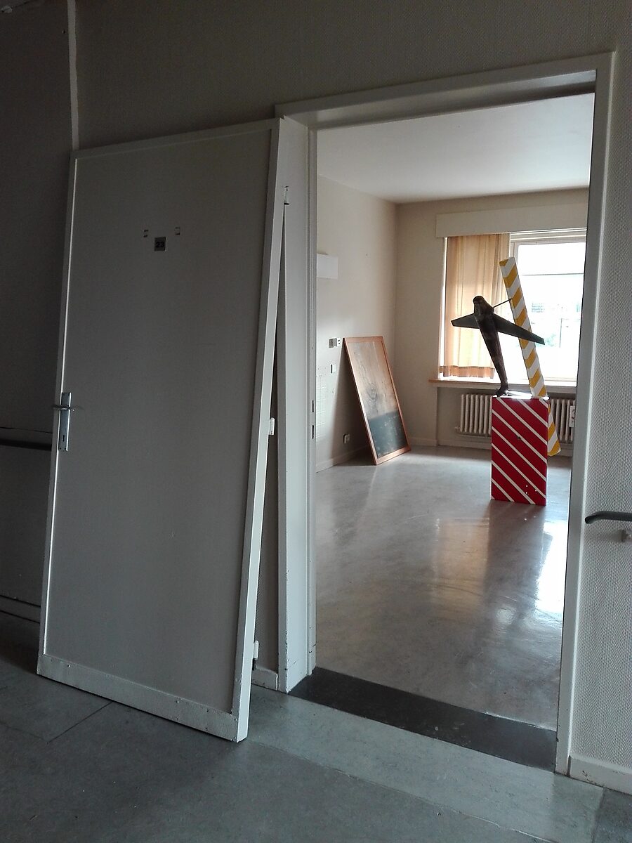 TarmaX: Exhibition view through an open doorway into a room with a metal airplane sculpture on a red-and-white striped pedestal; yellow-and-white diagonal beam behind it and painting leaning against the wall, Tarmac, Meerhout, 2017