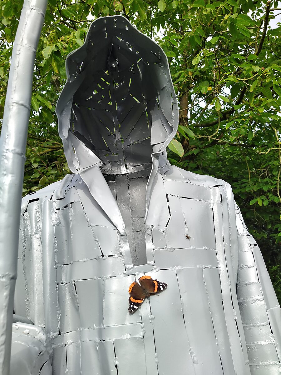 Interrupted Crossing — detail view of the grey-painted figure of Death with a butterfly resting on the chest of the sculpture.