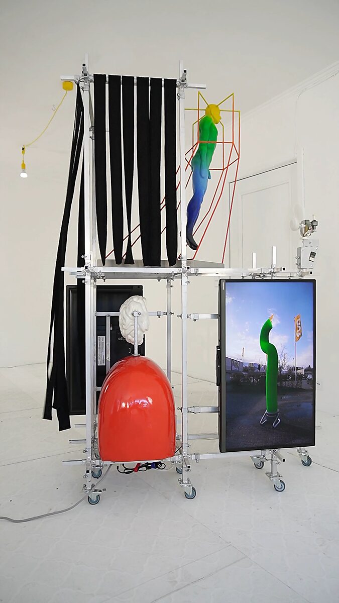 Thermiekbelofte — videostill van de installatie met het scherm waarop een skytube verschijnt, gecombineerd met het sculptuur Gedachtenkooi en bewegende stoffen linten die luchtstromen zichtbaar maken binnen het aluminium constructiesysteem.