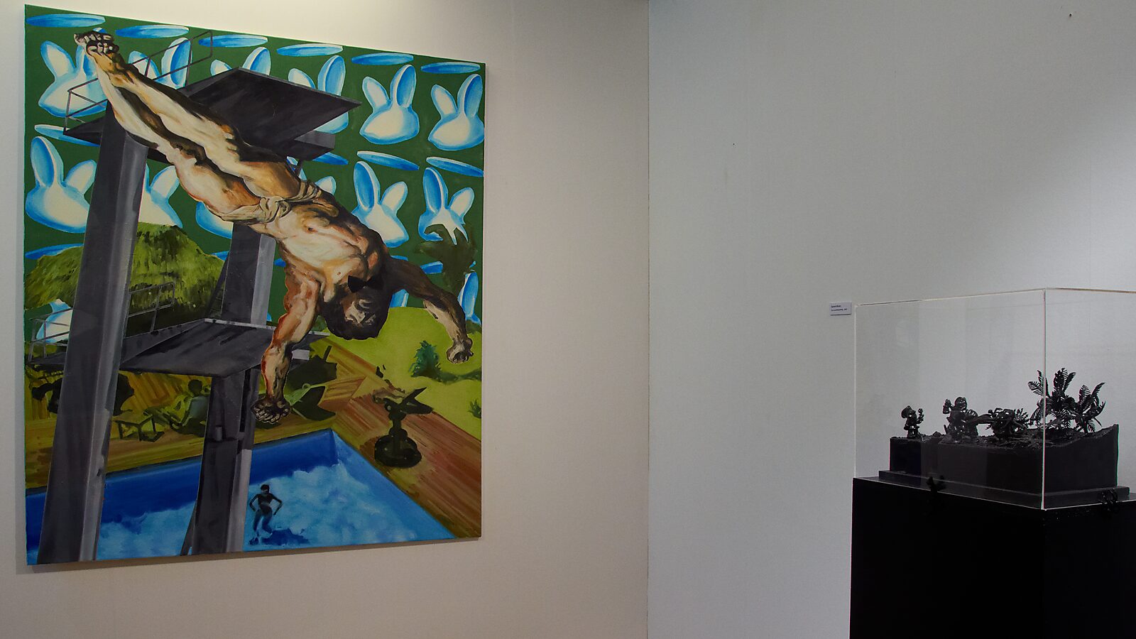 Duo exhibition with Raymond Minnen: Exhibition view with large figurative painting of a diving man against a green background with repeated white shapes; on the right a display case with small dark sculptures, CC De Kimpel, Alden Biesen, 2011