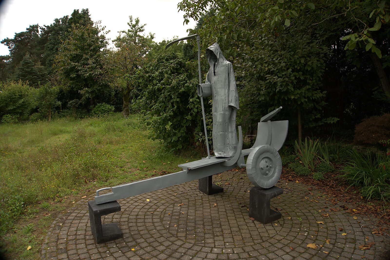 Interrupted Crossing — sculpture of Death standing on a boat trailer in a garden setting, finished with grey protective coating among trees and vegetation.