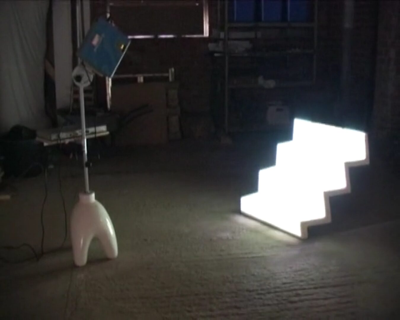Nu descendant par un ascenseur — videostill van een lichtgevende trapsgewijze sculptuur waarin een vrouwelijke figuur verschijnt als projectie, staand in een verticale nis die verwijst naar een dalende beweging.