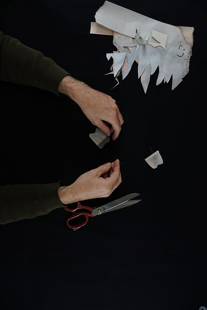Naming–Value Butterflies — process drawing: Hanging Man (1995) — hands place a small pin into a paper fragment while scissors and other leftover pieces lie on the table.