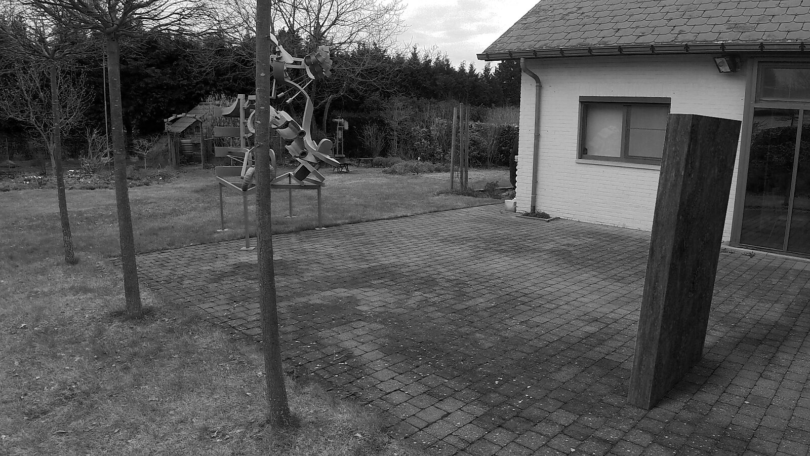 De klinker — videostill uit een 3D-animatie waarin een grote rechthoekige klinker rechtop op een terras verschijnt in een tuinomgeving.