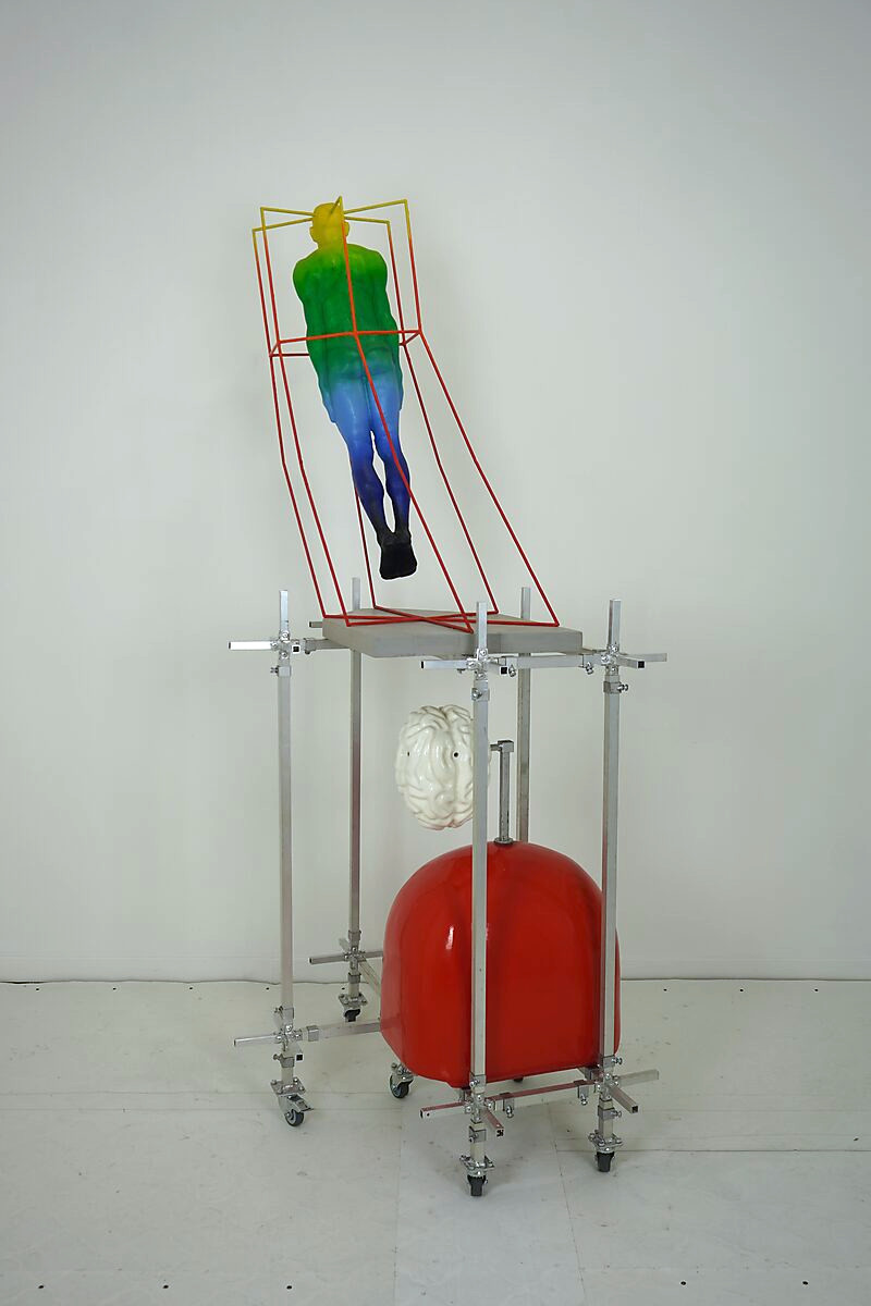 Gedachtenkooi — achteraanzicht van de sculptuur waarin het zwevende lichaam, de rode massa en het hersenelement samen een verticale relatie vormen tussen controle, gewicht en bewustzijn.