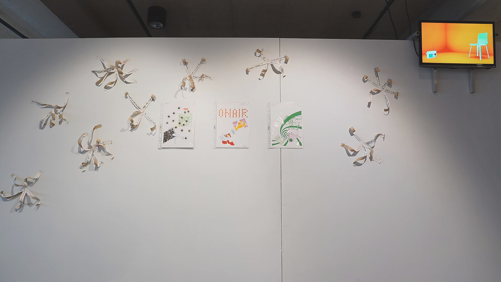 Transformatiestudio nr.1: warm of koud — tentoonstellingszicht: op een witte muur zijn meerdere lintvormige objecten uit Sfincterman bevestigd en als virusachtige vormen uitgespreid, gecombineerd met drie ingelijste Google-tekeningen; rechtsboven hangt ee