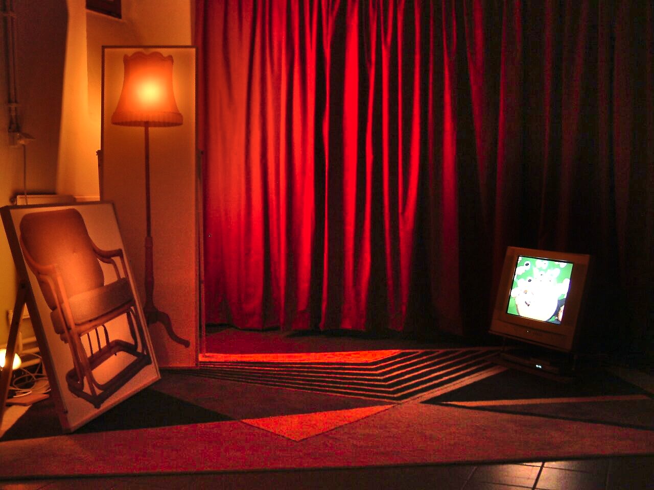 ‘Smaak’: Tentoonstellingszicht met rood verlichte gordijnen, staande lamp en kantelend beeld van een stoel; rechts een monitor met animatie, CC De Breughel, Bree, 2005
