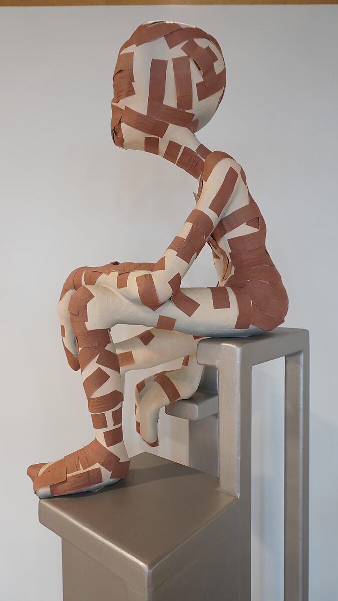 Alien Care, Bandage Phase 1 — rear view: alien figure with bandages seated sideways on the chair; back and rear of the head covered with bandages.