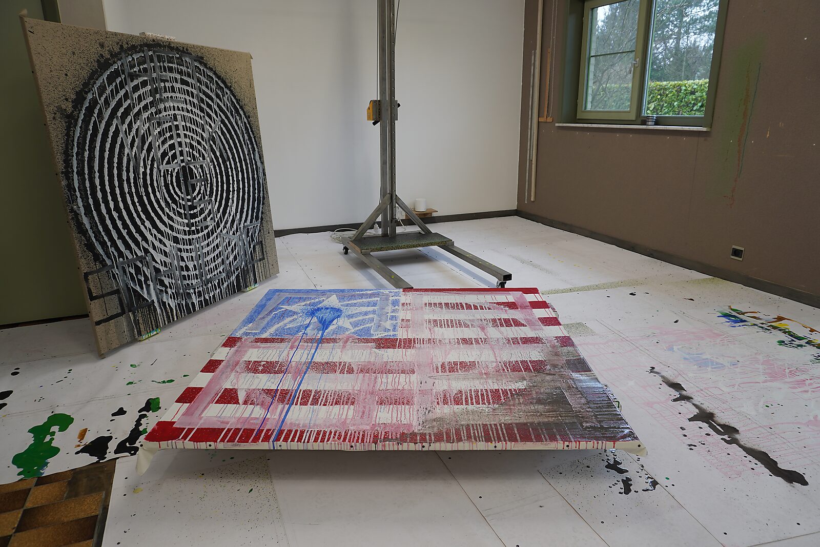 Doek met Amerikaanse vlagcompositie ligt op de vloer, met overschilderde lagen en druipsporen; op de achtergrond een ander doek met zwarte cirkelstructuur en een metalen constructie.
