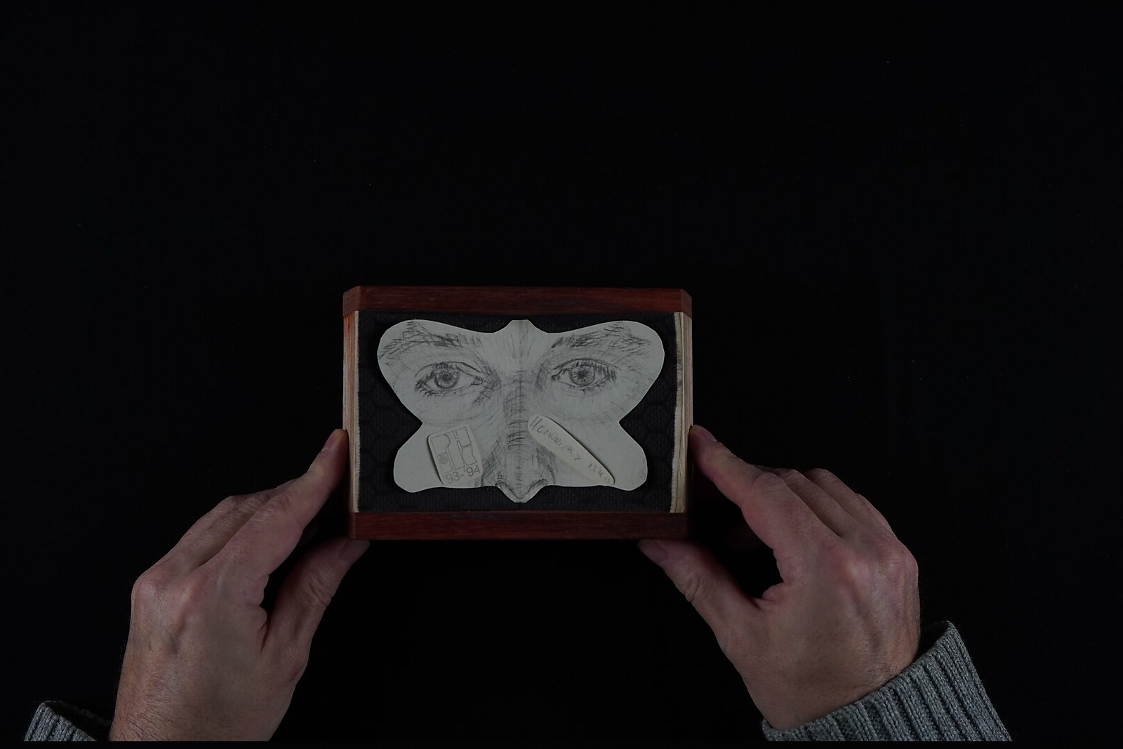 Naming–Value Butterflies — process drawing: study self-portrait 1 PHIKO (1993) — the finished butterfly-shaped drawing with eyes and nose lies in a small wooden box held by both hands.