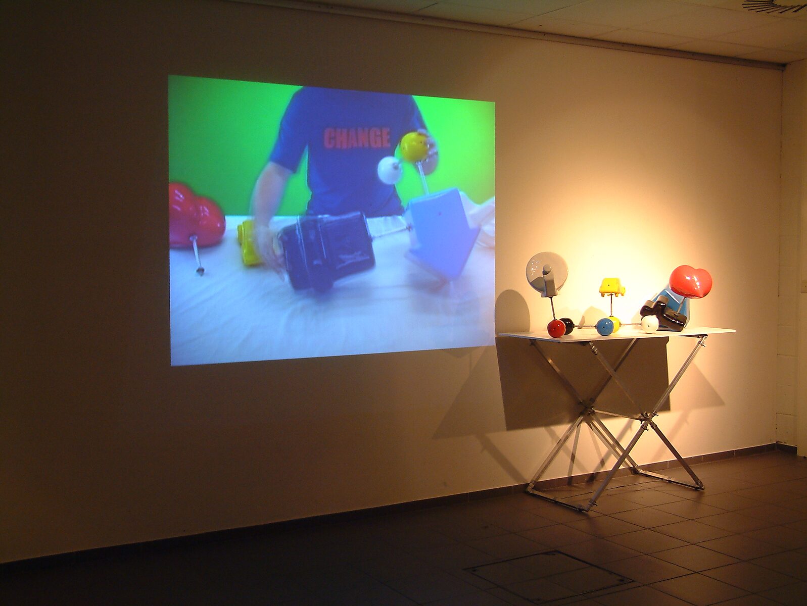 'Kies en verander project’: Tentoonstellingszicht met videoprojectie op de muur en tafel met kleurrijke objecten; metalen buisconstructies in de ruimte, CC De Kollebloem, Puurs, 2008.