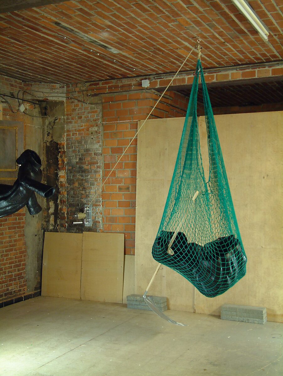 Doodsval nr. 2 — zwarte sculpturale figuur gevangen in een hangend groen net, zwevend boven de vloer terwijl een sikkelvormig element naar beneden wijst.