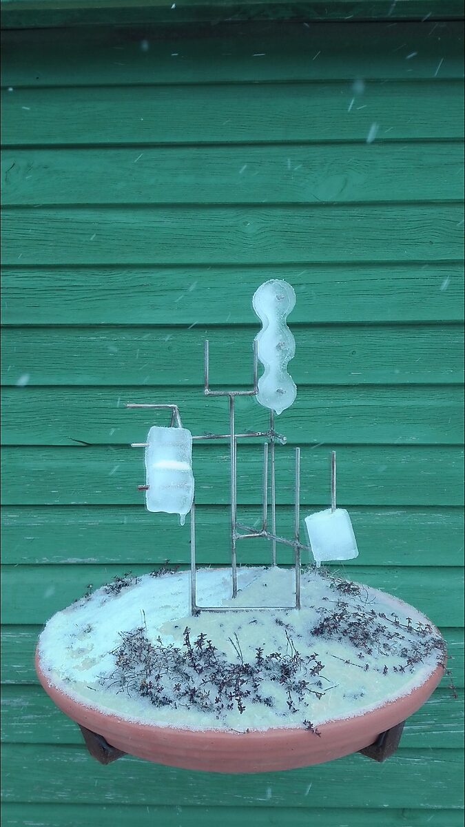 Entropische sculptuur nr. 1 — videostill van het metalen armatuur met ijsvormen, geplaatst op een schaal met aarde en sneeuw voor een groene houten wand, opgenomen in de winter.