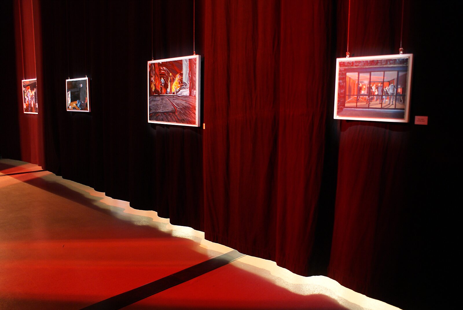 ‘The Modern Cavemen’: Exhibition view with suspended oil paintings in front of black curtains; intense red lighting illuminates the space, The Modern Cavemen, Dommelhof, Pelt, 2012.