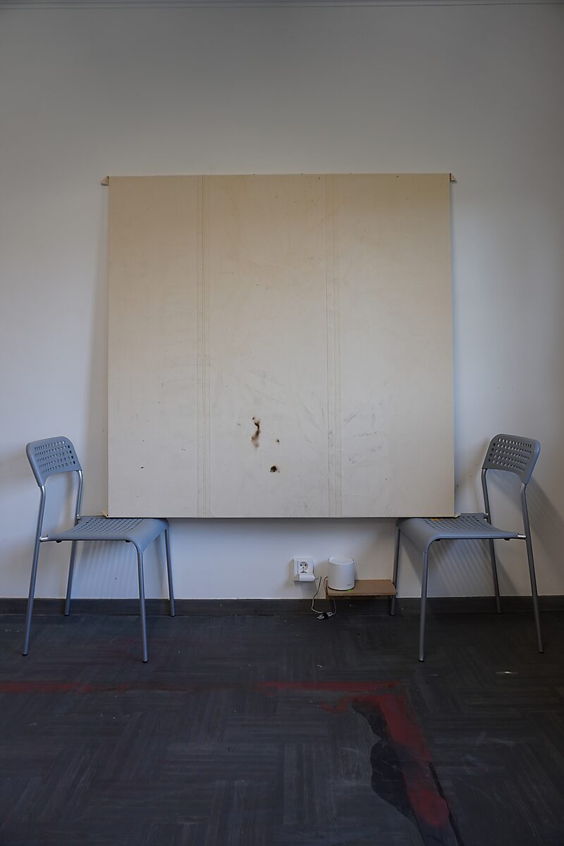 Leeg groot paneel met sporen van gebruik en vlekken, opgesteld tussen twee stoelen; rode verfsporen zichtbaar op de vloer ervoor.