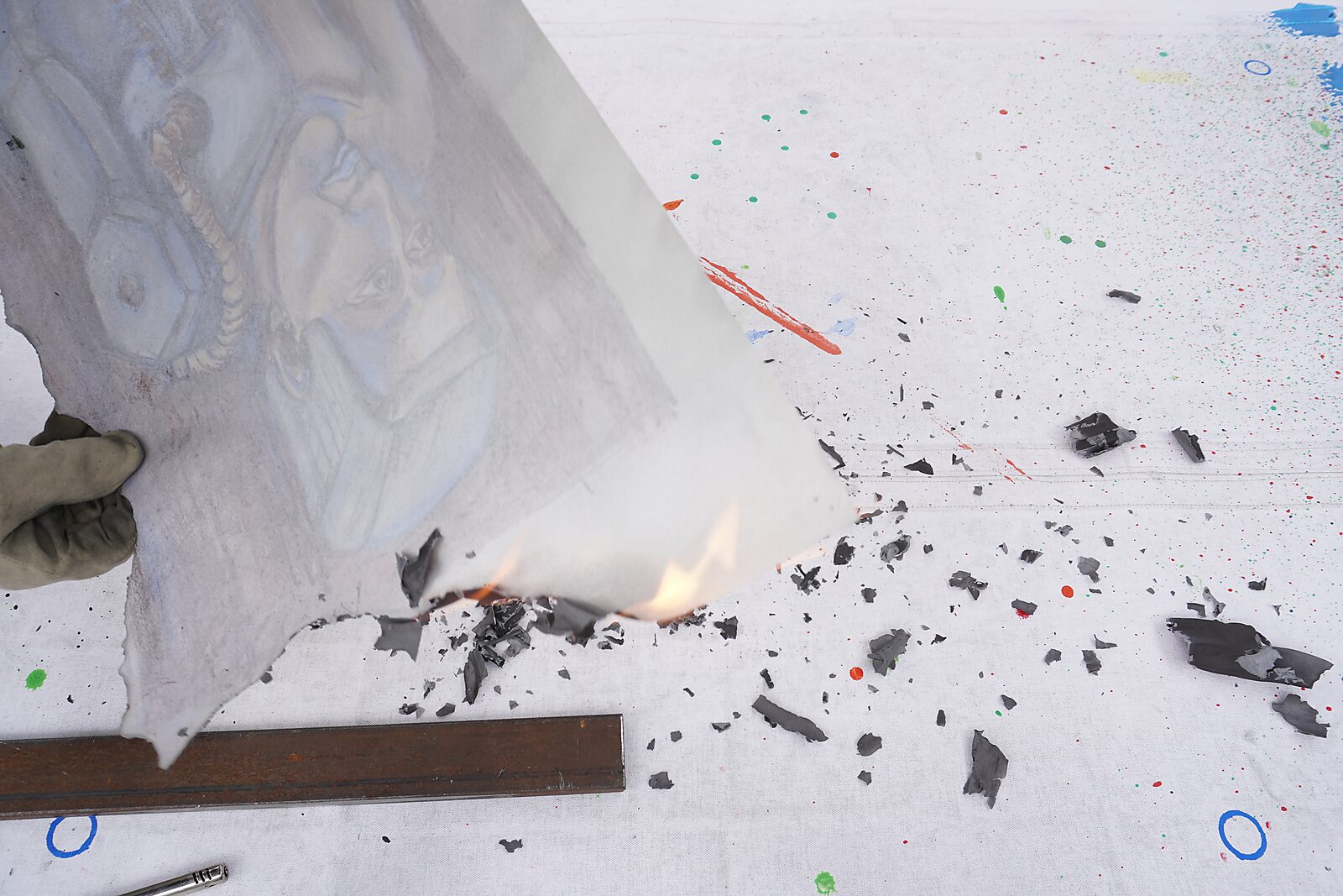 The Divider — process photo: edges of a drawing being burned away with fire above a paint-splattered work surface.