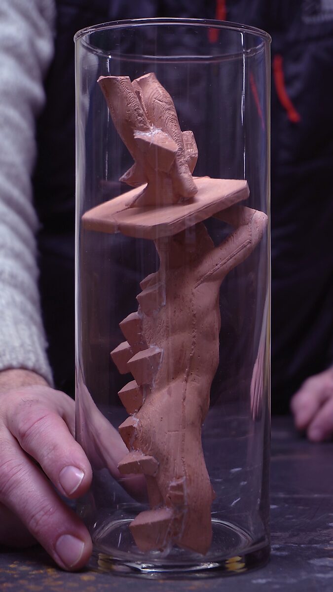 Gebroken volume — videostill van het beeldje dat in stukken wordt gebroken en in de glazen cilinder kantelt.
