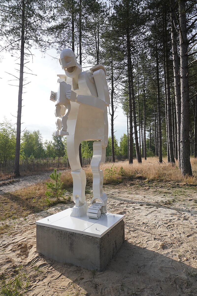 Saturnus — witte sculptuur van een gestileerde menselijke figuur die voorovergebogen een kleiner figuur vasthoudt, geplaatst op een betonnen sokkel in het zandlandschap van de Sahara in Lommel, omringd door dennenbos.