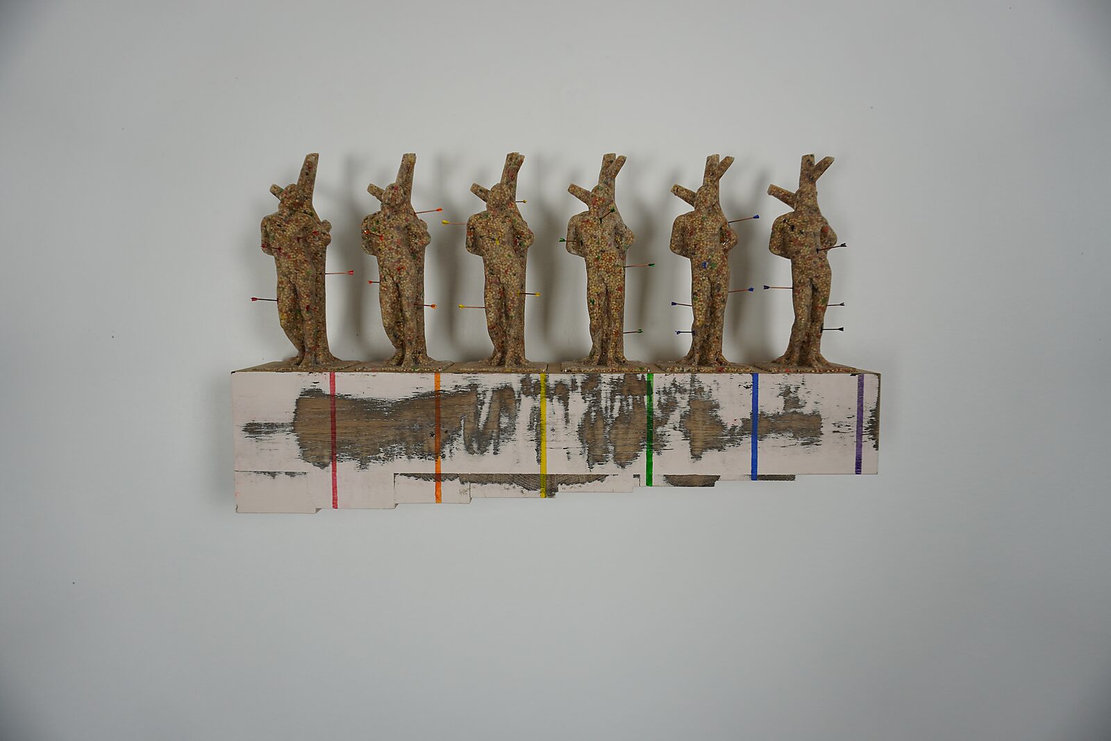 Saint Sebastian in LGBTQ+ style — sculpture with six identical casts from the same mold, pierced by arrows, on a wooden base with rainbow-colored vertical stripes.