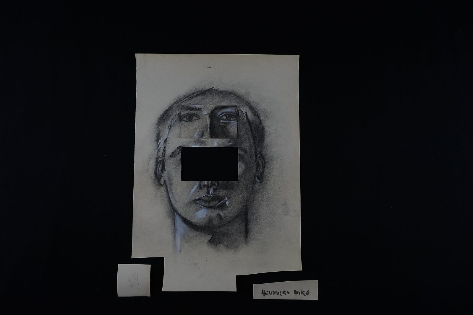Benoemens-waardvlinders — Naming–Value Butterflies — proces tekening: studie zelfportret 2 PHIKO (1993), Gevangen (2025) — het portret met een rechthoekige opening in het gezicht ligt op tafel met losse naam- en labelstroken eronder.