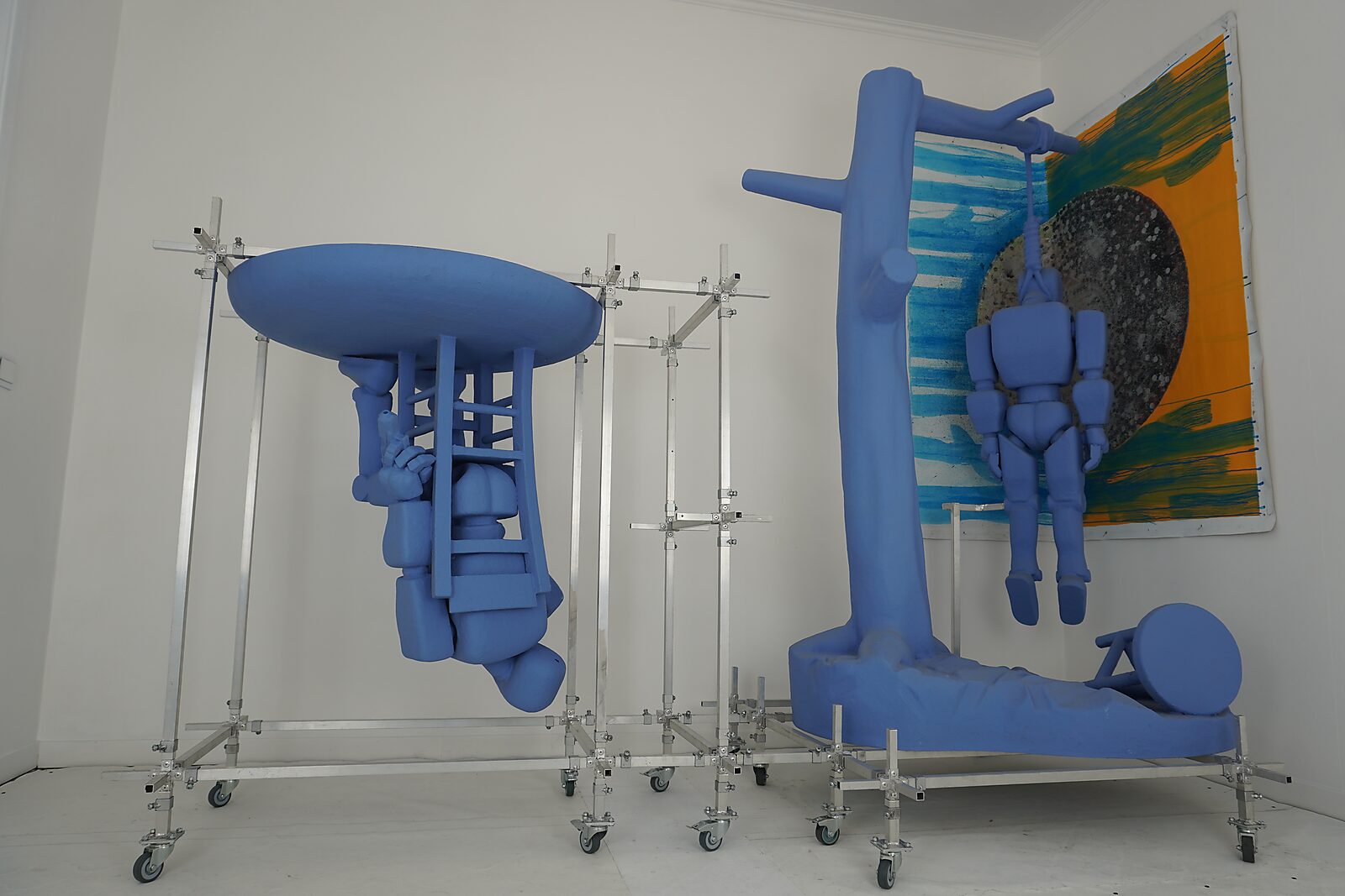Twee blauwe sculpturale constructies binnen een aluminium framesysteem; één figuur hangt aan een structuur terwijl een andere ondersteboven aan een platform is bevestigd, tegen een witte muur met schilderij.