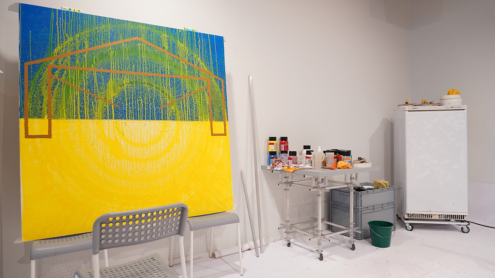 Blue-yellow painting with concentric circles and vertical drips, overlaid with an orange geometric shape; in front a chair and next to it a worktable with paint jars and materials.