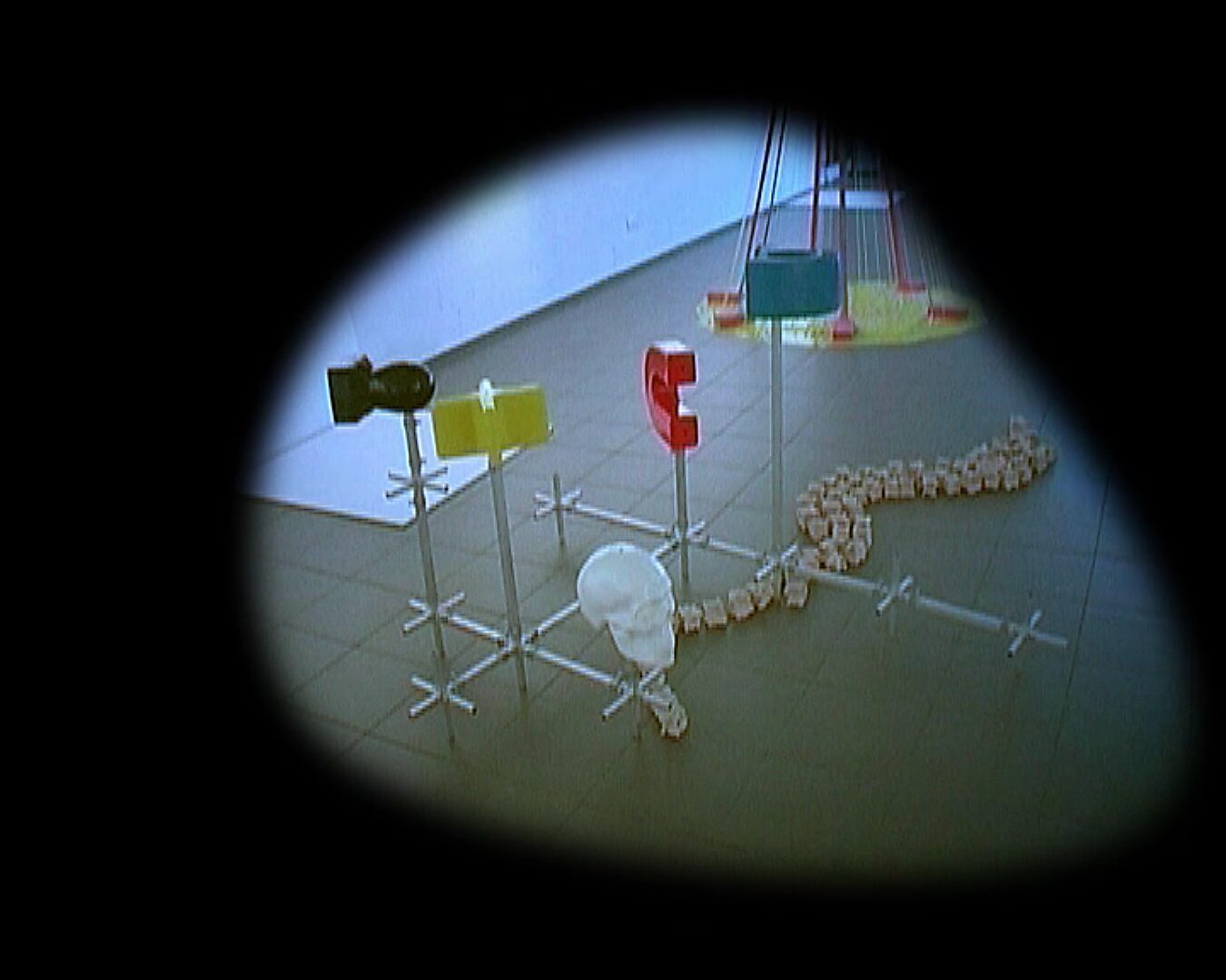 Kies en verander, Installatie in beweging — videostill: modulaire opstelling met gekleurde elementen en een schedelvorm, verbonden door metalen stangen, in een tentoonstellingsruimte.