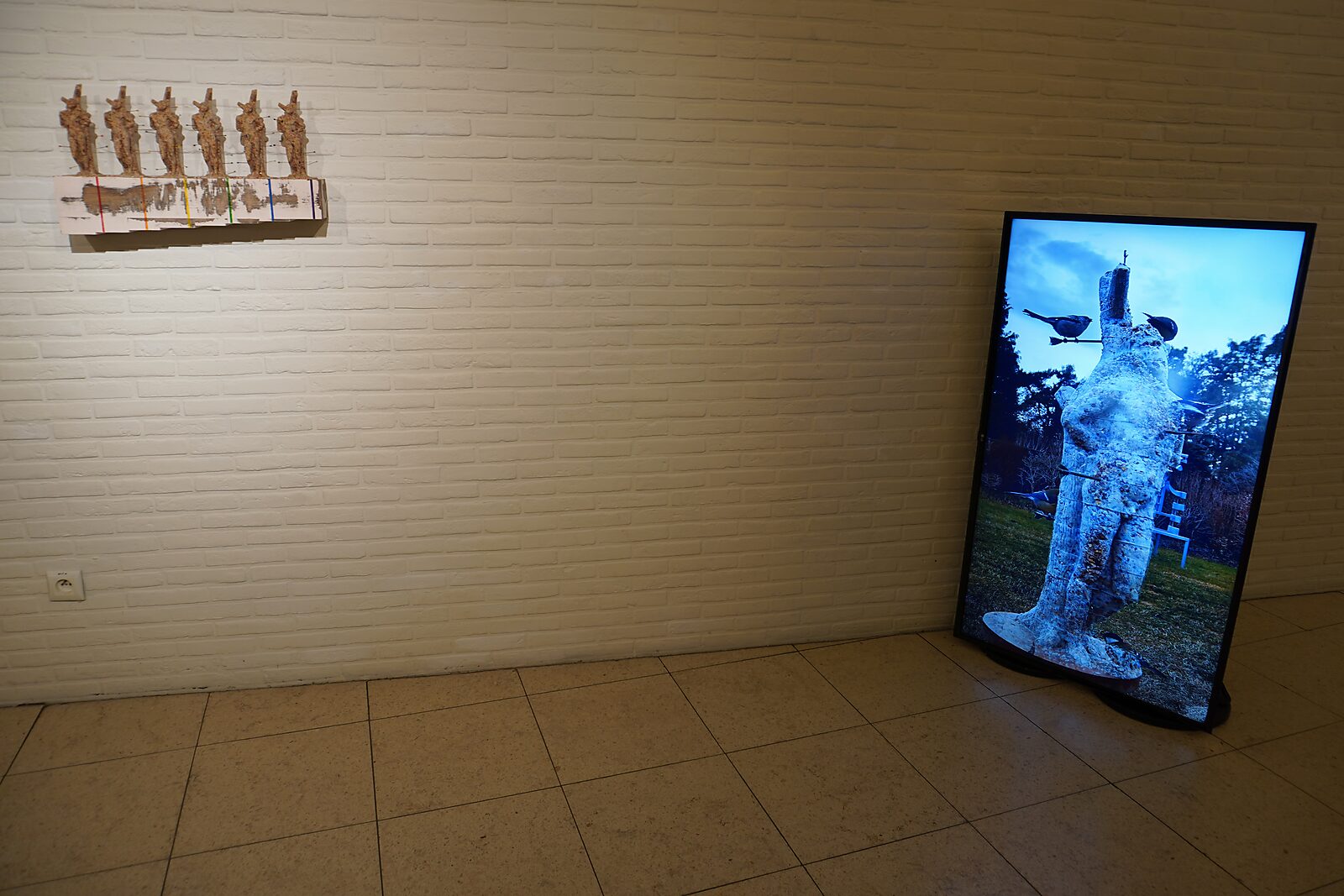 'Sfincterman for President’: Exhibition view with a wall piece (row of small forms) and a vertical video screen showing an outdoor figure with birds, Dommelhof, Pelt, 2024.