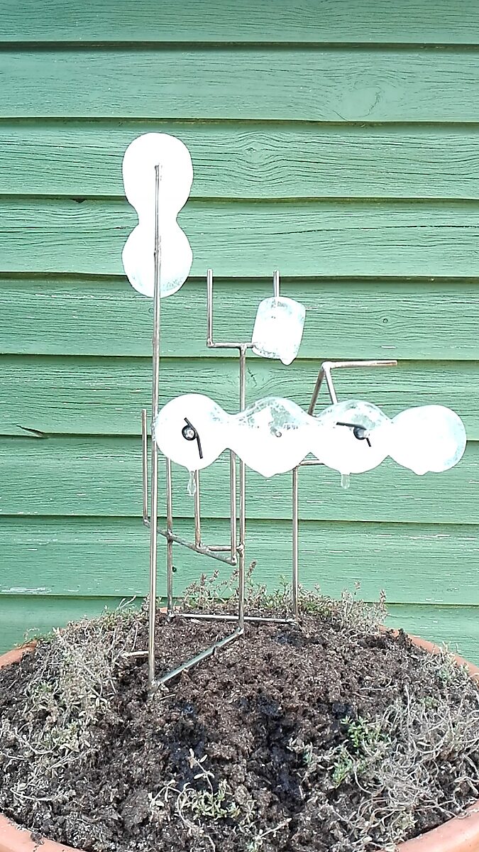 Entropic Sculpture No. 1 — videostill of a metal armature with ice forms attached, placed on a pot of soil in front of a green wooden wall, recorded in summer.