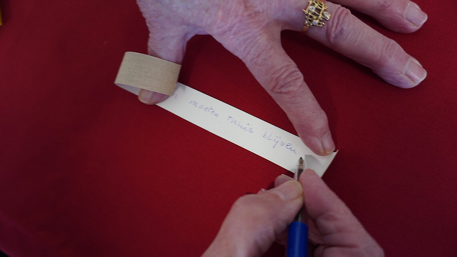 Sfincterman nr.0.0 — videostill handeling: een hand schrijft met een blauwe pen tekst op een smalle papieren strook die op een rode ondergrond ligt, terwijl een stuk tape rond een vinger zit.