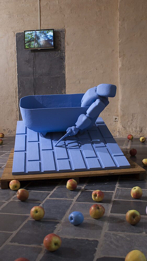 ‘Strange Fruit’: Blauwe robotfiguur half liggend in een badkuip op een hellend platform; echte appels verspreid over de vloer, videoscherm en schilderijen op de achtergrond, Gasthuiskapel, Borgloon, 2015.