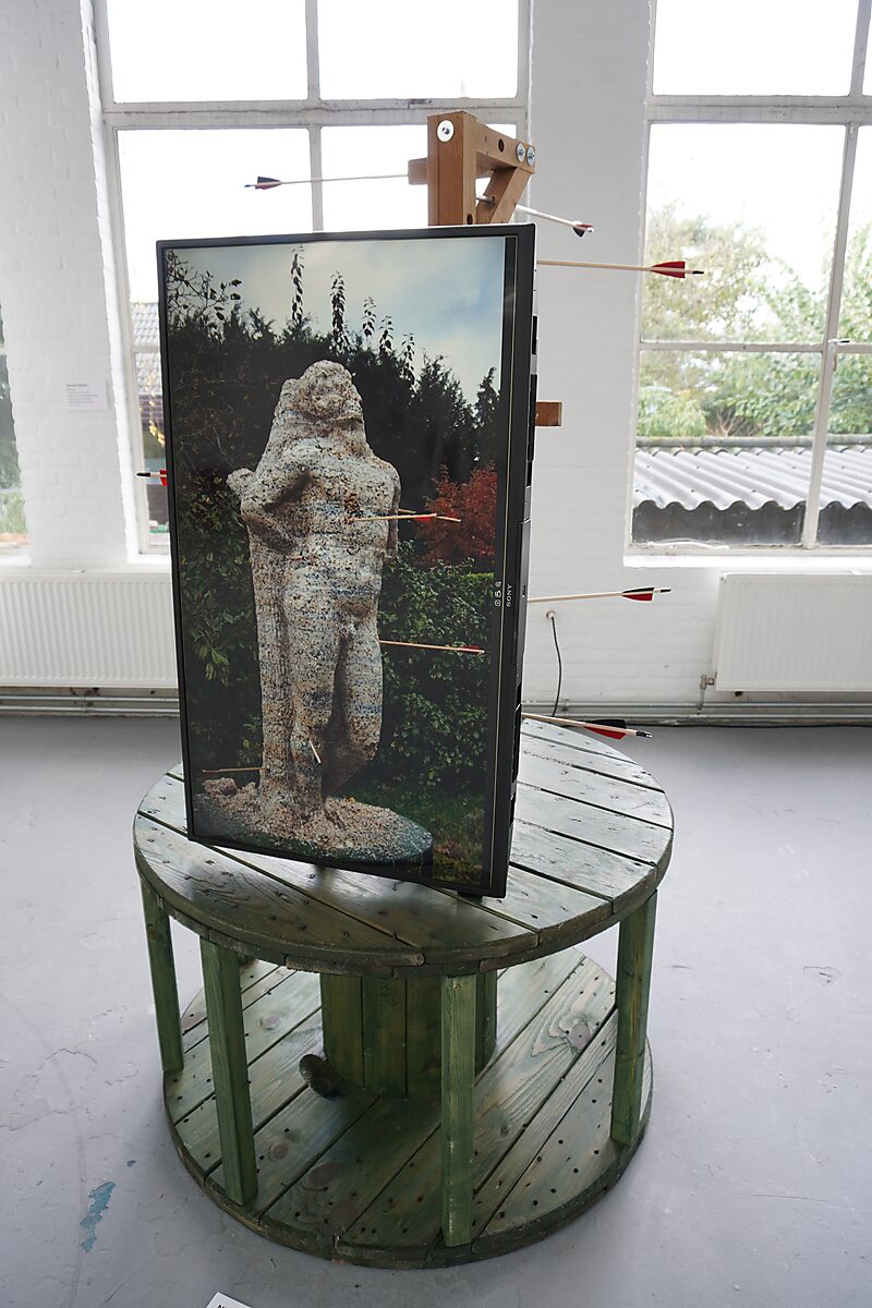 ‘Malabou in Malibu’: Beeldscherm met foto van dezelfde doorboorde figuur, vastgezet met pijlen; scherm op ronde houten kabelhaspel voor groot raam, De Fabriek, Eindhoven, 2023.