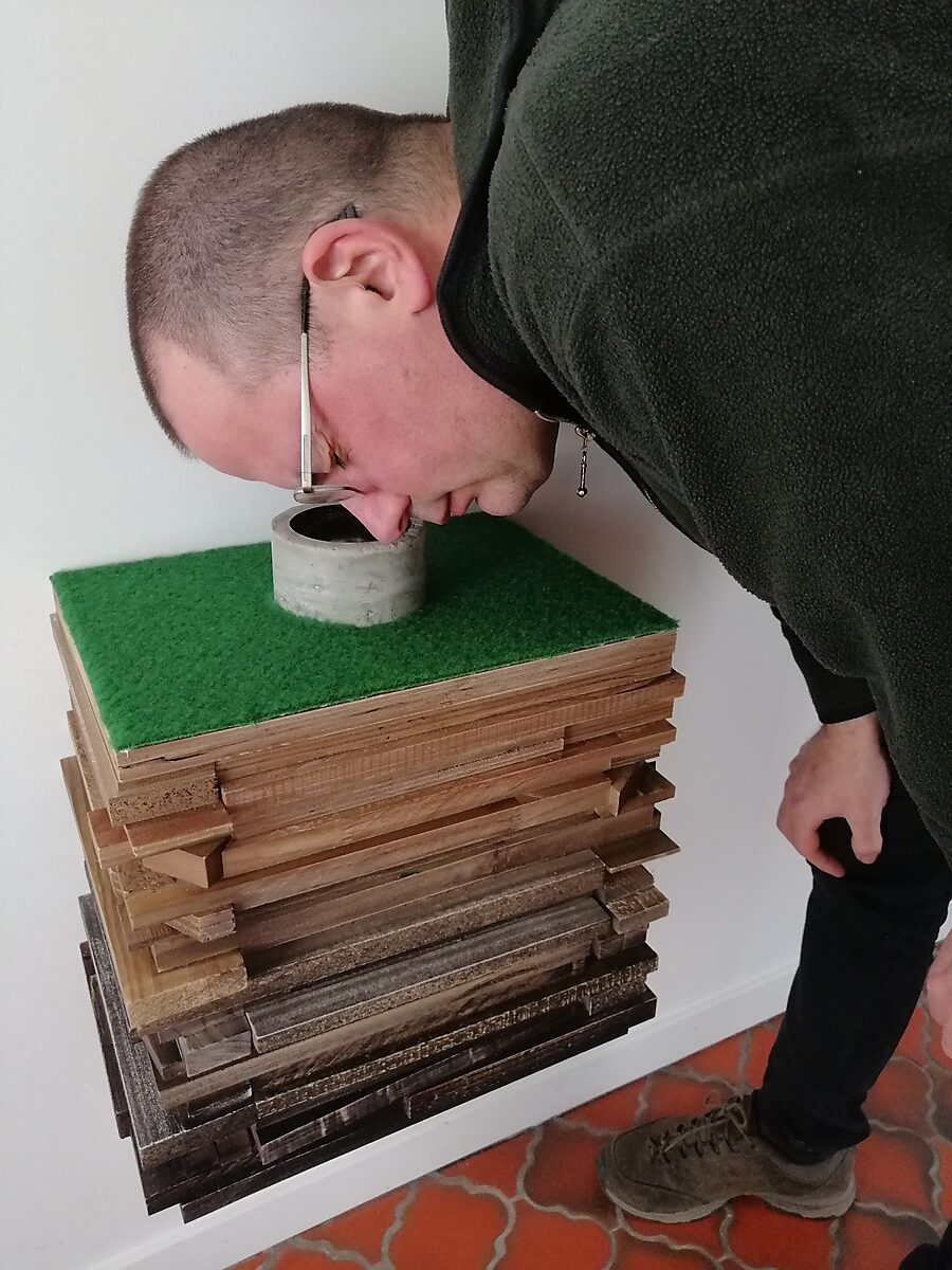 Put van fluïditeiten — bezoeker die zich voorover buigt om in de betonnen putopening bovenop het wandsculptuur te kijken.