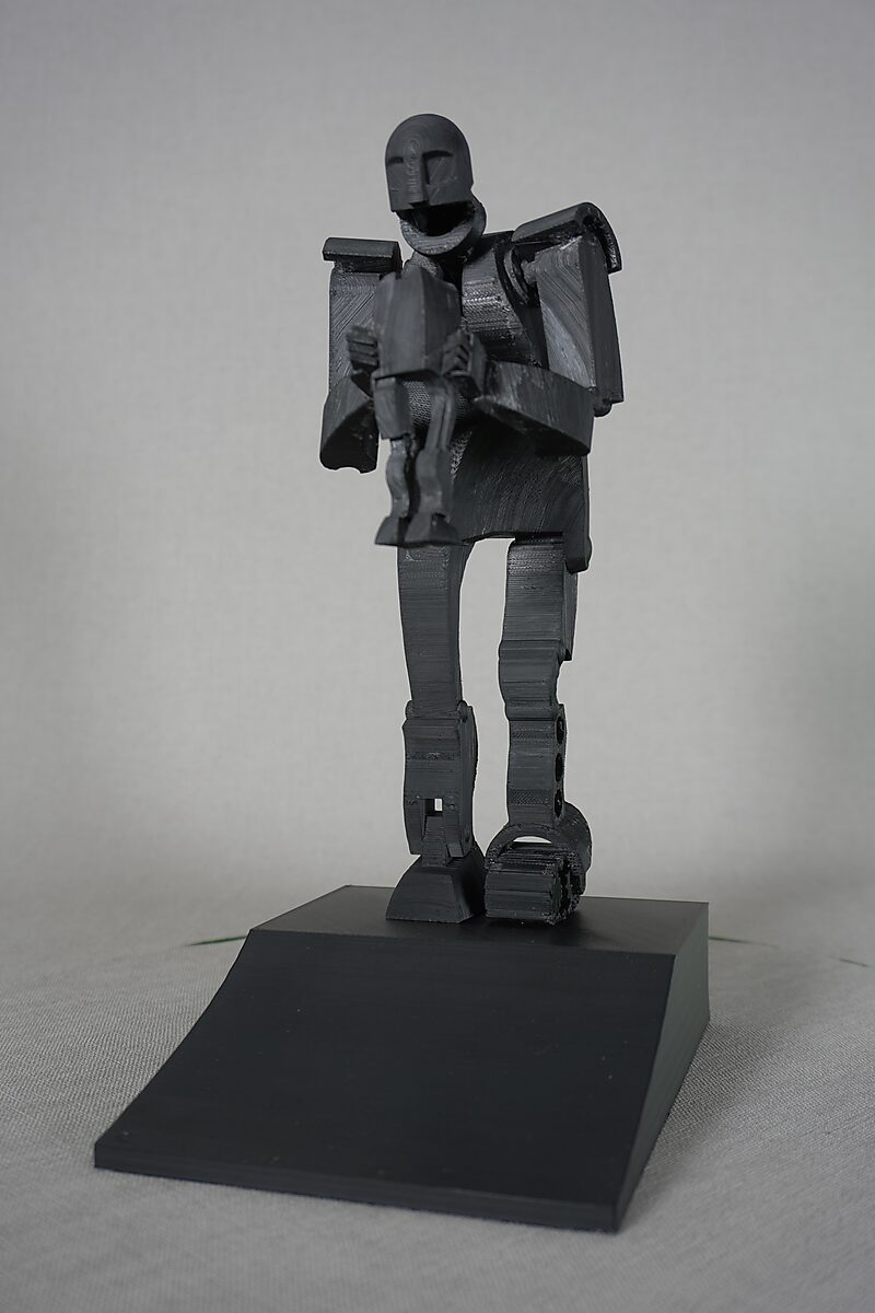 Saturn — black 3D-printed miniature in frontal view of a stylized figure presenting a smaller figure, placed on a black pedestal.