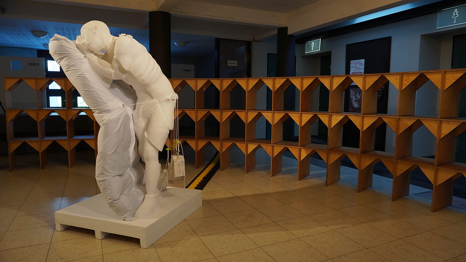 'Sfincterman for President’: Exhibition view with a white figure sculpture leaning against a large pillow on a plinth; wooden structure in the background, Dommelhof, Pelt, 2024.