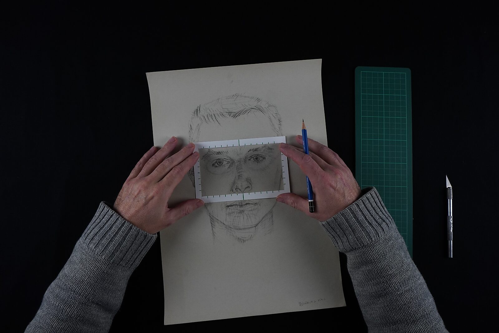 Naming–Value Butterflies — process drawing: study self-portrait 1 PHIKO (1993) — two hands hold a small drawn fragment with eyes above a larger self-portrait; a pencil, cutting knife and cutting mat lie on the table.