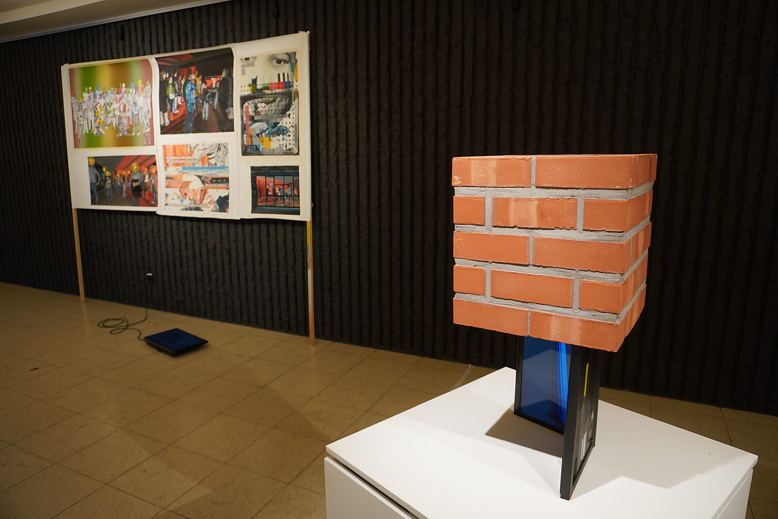 'Sfincterman for President’: Tentoonstellingszicht met baksteenblok op sokkel; op de achtergrond een bord met beeldcollage en een klein scherm op de vloer, Dommelhof, Pelt, 2024.