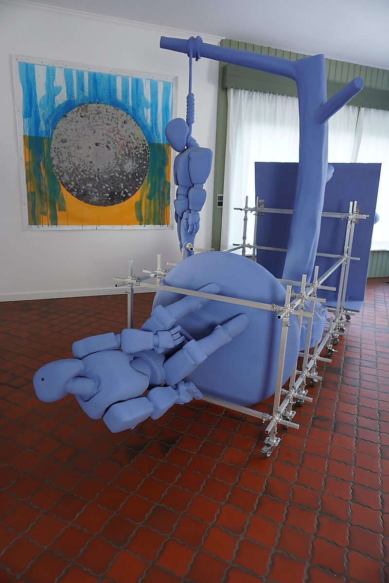 Blue sculptural figures and forms mounted in an aluminum construction system on wheels; one figure hangs from a beam, another lies bent over a rounded shape, with a painting in the background.