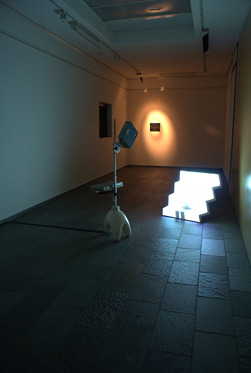 Nu descendant par un ascenseur — installation view at CC De Adelberg in Lommel showing a video projector on a stand aimed at a luminous stepped sculpture placed on the floor in a darkened exhibition space.