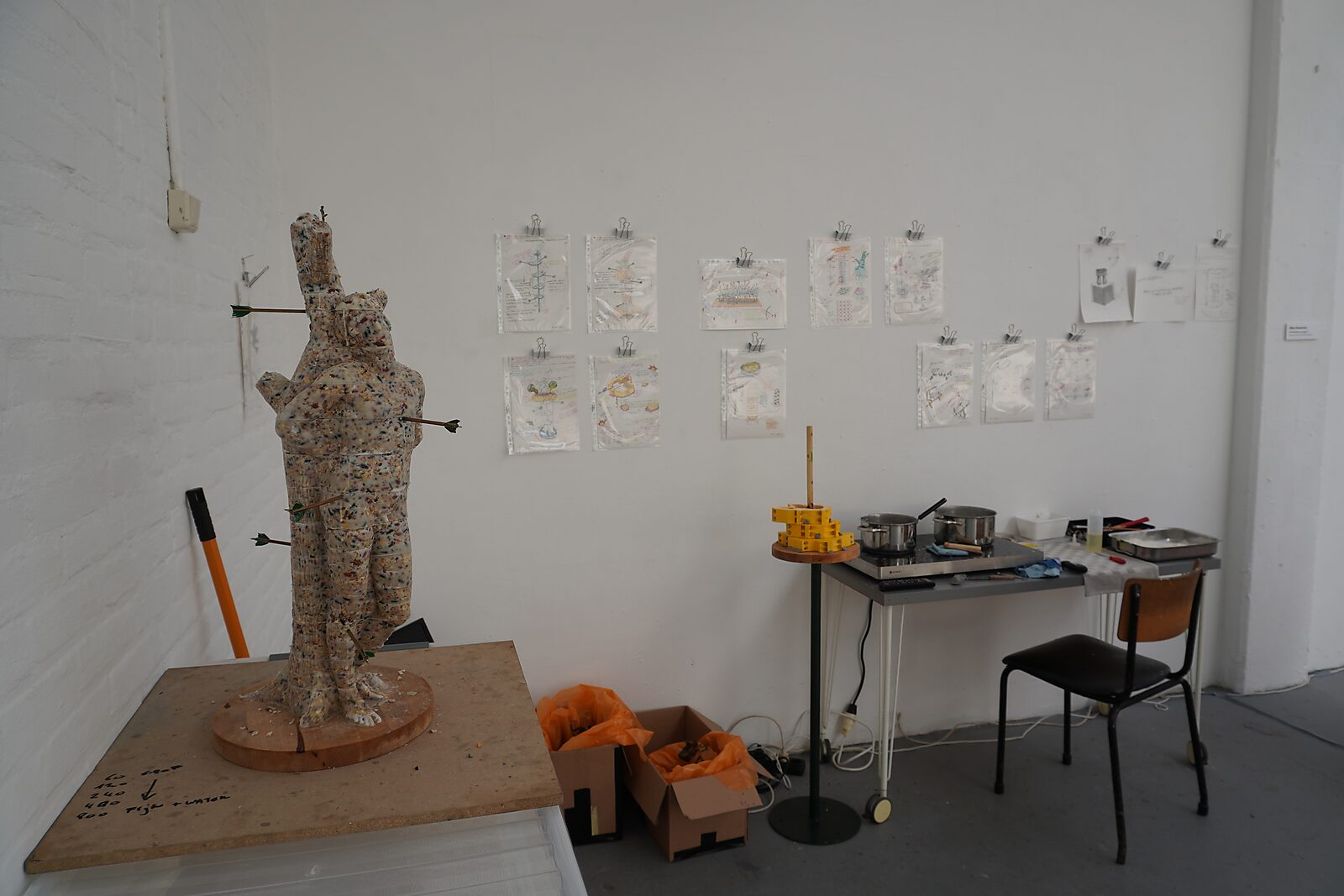 ‘Malabou in Malibu’: Small human figure in speckled material pierced with arrows on a wooden turntable; study drawings and worktable in the background, De Fabriek, Eindhoven, 2023.