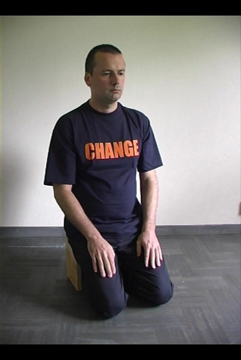 Kies en verander nr. 2 — videostill van de kunstenaar knielend op de vloer, met open ogen en het woord “CHANGE” op het T-shirt.