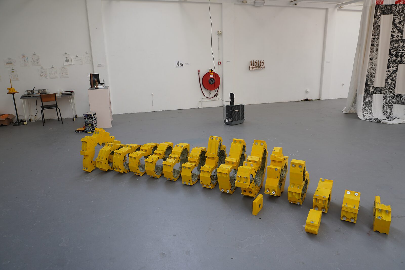 ‘Malabou in Malibu’: Long row of bright yellow industrial segments arranged on the floor in a white exhibition space; worktable and small sculptures against the wall, De Fabriek, Eindhoven, 2023.