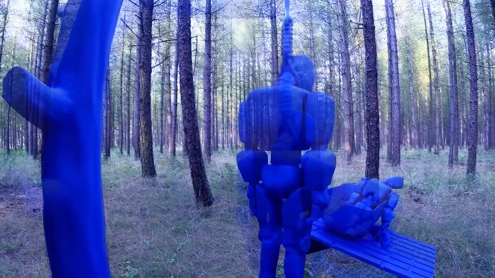 Imaginary Landscapes — video still showing robot figures reappearing in a forest environment; one figure hangs from a structure while another body rests on a bench, turning the landscape into a site of observation and renewal.