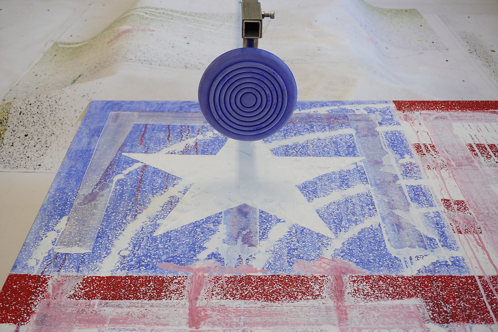 Close-up of a red-white-blue canvas based on the American flag, featuring a star-shaped stencil and a circular stamp-like form hovering above the surface; spray mist and drips are visible.